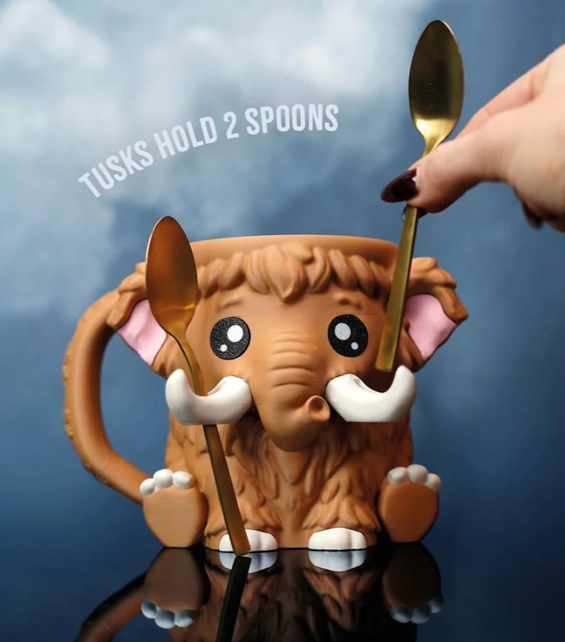 May include: A brown ceramic mug shaped like a cartoon mammoth with a trunk, tusks, and furry details. The mug has a handle and holds two gold spoons. Text on the image reads "TUSKS HOLD 2 SPOONS".