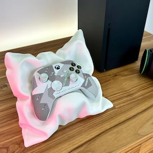 May include: A white and black Xbox controller with green buttons and the text "Project Scarlett" on the front. The controller is resting on a white pillow on a wooden surface.