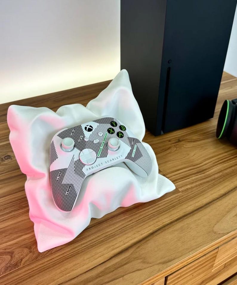 May include: A white and black Xbox controller with green buttons and the text "Project Scarlett" on the front. The controller is resting on a white pillow on a wooden surface.