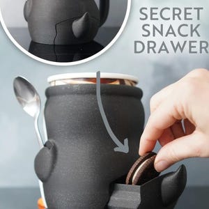 May include: Black ceramic snack container with a secret drawer, a spoon, and the text "SECRET SNACK DRAWER". A hand is reaching into the drawer to grab a cookie. The container has a unique, cartoonish design.