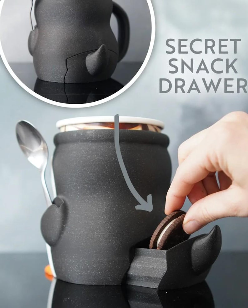 May include: Black ceramic snack container with a secret drawer, a spoon, and the text "SECRET SNACK DRAWER". A hand is reaching into the drawer to grab a cookie. The container has a unique, cartoonish design.