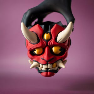 May include: A red and black Oni mask with white horns and fangs, and yellow eyes. The mask is held by a gloved hand against a purple background. The mask is a traditional Japanese demon mask.