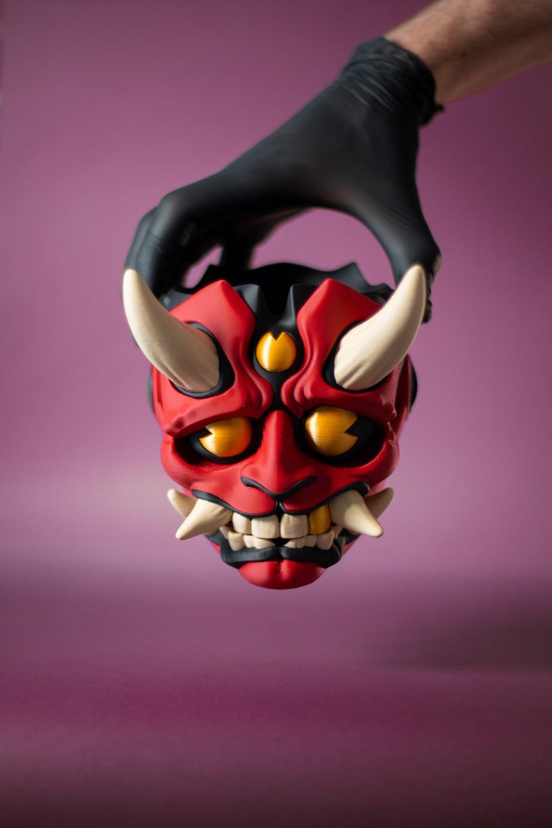 May include: A red and black Oni mask with white horns and fangs, and yellow eyes. The mask is held by a gloved hand against a purple background. The mask is a traditional Japanese demon mask.