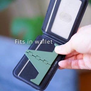 Może przedstawiać: Czarny, dwukrotnie składany portfel z zielonym stojakiem na telefon. Portfel ma przezroczyste okienko na dow&oacute;d i białe paski. Widoczny jest napis "Fits in wallet". Stojak na telefon jest złożony.