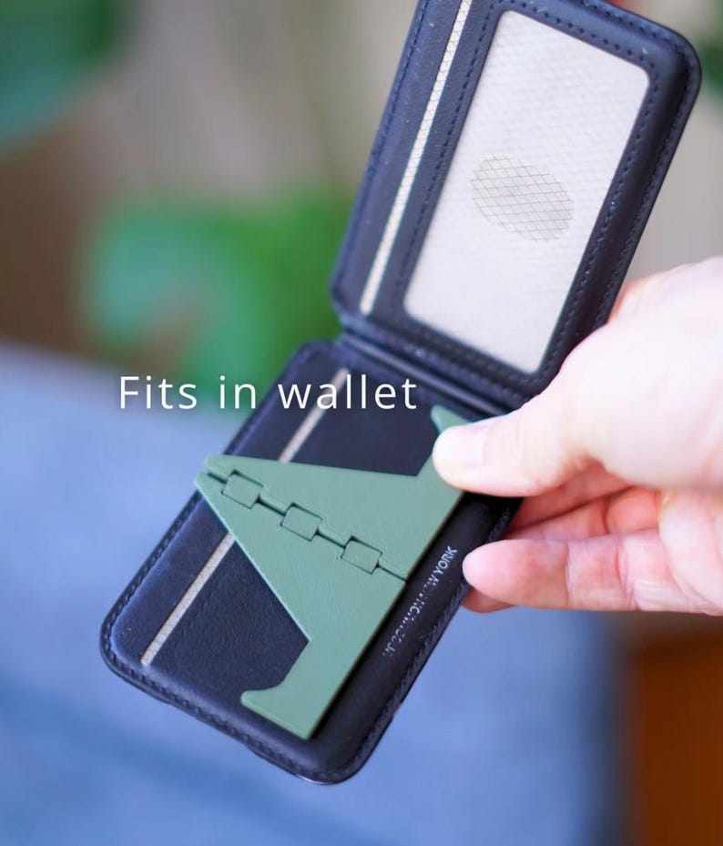 Może przedstawiać: Czarny, dwukrotnie składany portfel z zielonym stojakiem na telefon. Portfel ma przezroczyste okienko na dow&oacute;d i białe paski. Widoczny jest napis "Fits in wallet". Stojak na telefon jest złożony.
