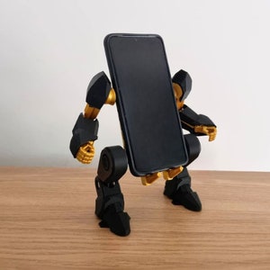 May include: A black and gold robot phone stand with articulated arms and legs. The stand is holding a black smartphone.