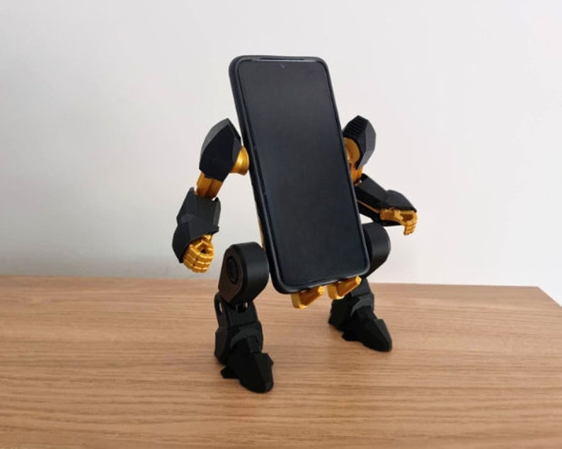 May include: A black and gold robot phone stand with articulated arms and legs. The stand is holding a black smartphone.