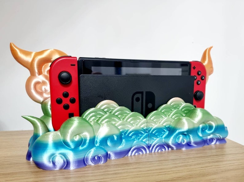 May include: A 3D printed Nintendo Switch stand with a colorful cloud design. The stand features two horns on either side and a slot for the Switch console.