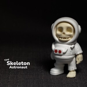 May include: A white skeleton astronaut figurine with red details on the suit. The figurine is wearing a white helmet with a clear visor. The figurine is standing on a black surface. Tiny Skeleton Astronaut.