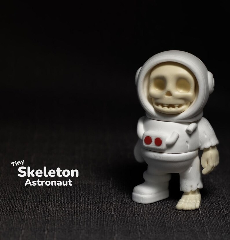 May include: A white skeleton astronaut figurine with red details on the suit. The figurine is wearing a white helmet with a clear visor. The figurine is standing on a black surface. Tiny Skeleton Astronaut.