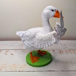 May include: A white goose figurine with orange beak and feet, standing on a green circular base. The goose is sculpted with detailed feathers and is posed with one wing raised. The figurine is approximately 20 cm tall.