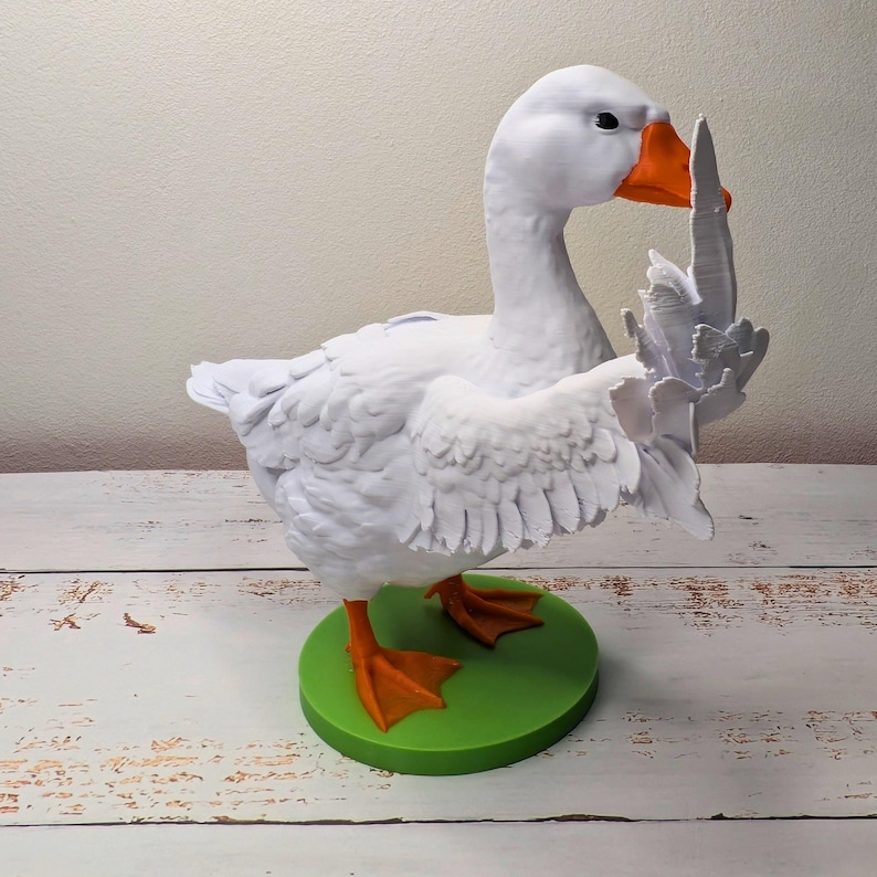 May include: A white goose figurine with orange beak and feet, standing on a green circular base. The goose is sculpted with detailed feathers and is posed with one wing raised. The figurine is approximately 20 cm tall.