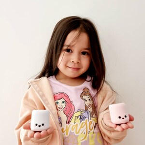 Op de afbeelding: Een jong meisje draagt een roze shirt met bedrukte Disney-prinsessen en houdt twee kleine, witte en roze marshmallow-vormige speelgoedjes vast. De speelgoedjes hebben vrolijke gezichten erop getekend.