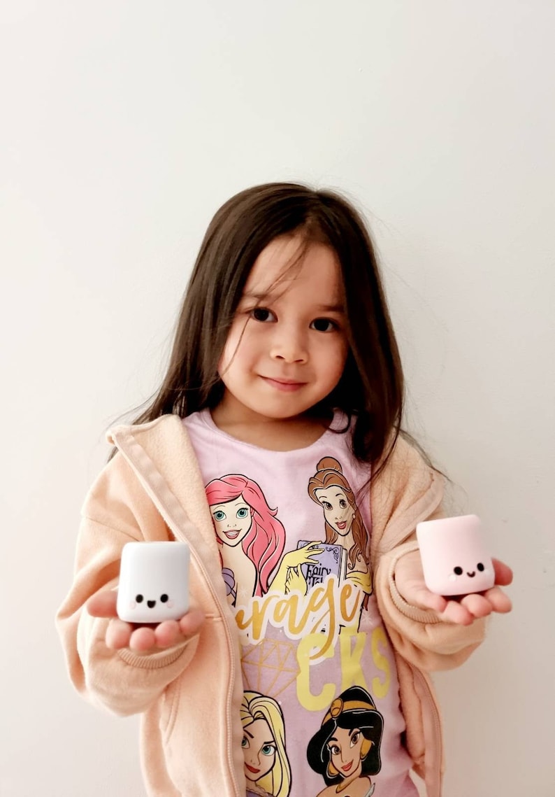 Op de afbeelding: Een jong meisje draagt een roze shirt met bedrukte Disney-prinsessen en houdt twee kleine, witte en roze marshmallow-vormige speelgoedjes vast. De speelgoedjes hebben vrolijke gezichten erop getekend.