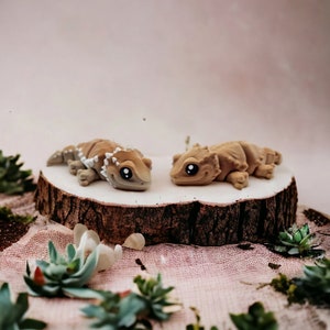 May include: Two small, brown, ceramic figurines of bearded dragons on a wooden slice. The figurines have large, black eyes and are facing each other.