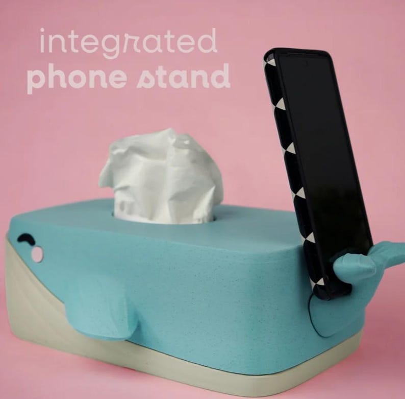 May include: A light blue whale-shaped tissue box with an integrated phone stand. The tissue box has a white tissue sticking out of the top. The phone stand is black and holds a black phone. The text "integrated phone stand" is above the tissue box.