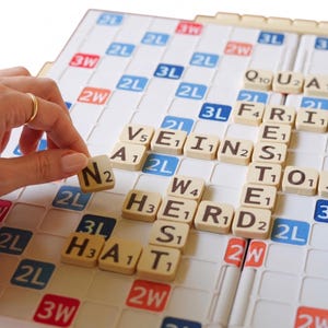 Puede incluir: Un tablero de Scrabble con fichas de letras beige formando palabras. Una mano con un anillo dorado coloca una ficha "N" en el tablero. El tablero tiene casillas azules, rojas y blancas con multiplicadores de puntos.