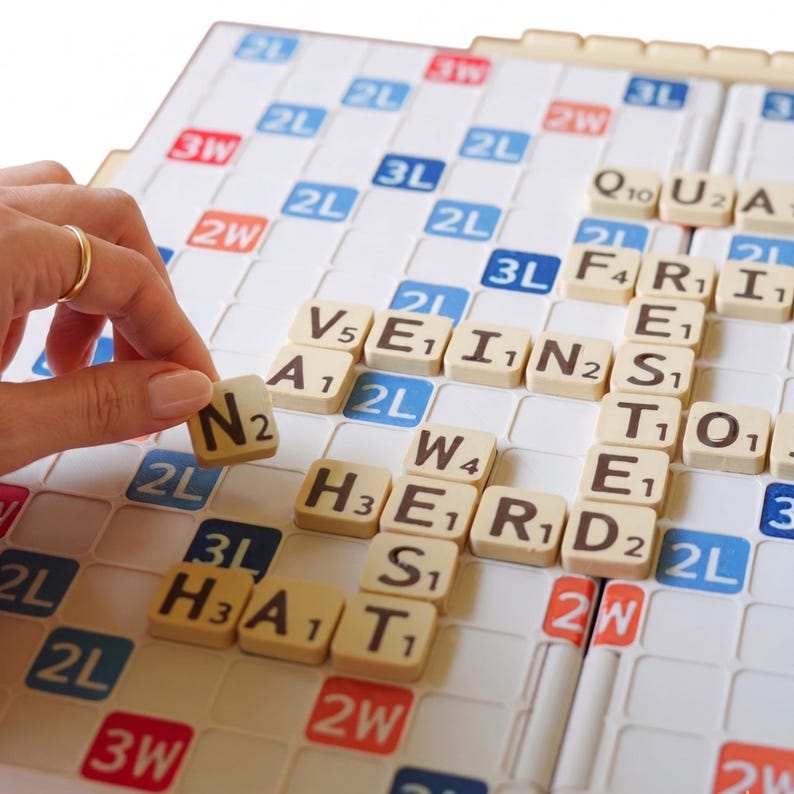 Puede incluir: Un tablero de Scrabble con fichas de letras beige formando palabras. Una mano con un anillo dorado coloca una ficha "N" en el tablero. El tablero tiene casillas azules, rojas y blancas con multiplicadores de puntos.