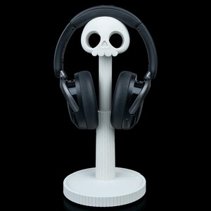 May include: A white skull-shaped headphone stand with a round base and a vertical support. Black headphones rest on the stand, their earcups facing forward. The stand is set against a black background.