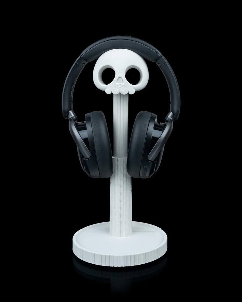 May include: A white skull-shaped headphone stand with a round base and a vertical support. Black headphones rest on the stand, their earcups facing forward. The stand is set against a black background.