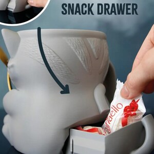 May include: A gray cat-shaped mug with a secret snack drawer. The mug has a handle and a textured design on the back. The words "SECRET SNACK DRAWER" are printed above the mug. A hand is placing a wrapped candy into the drawer.