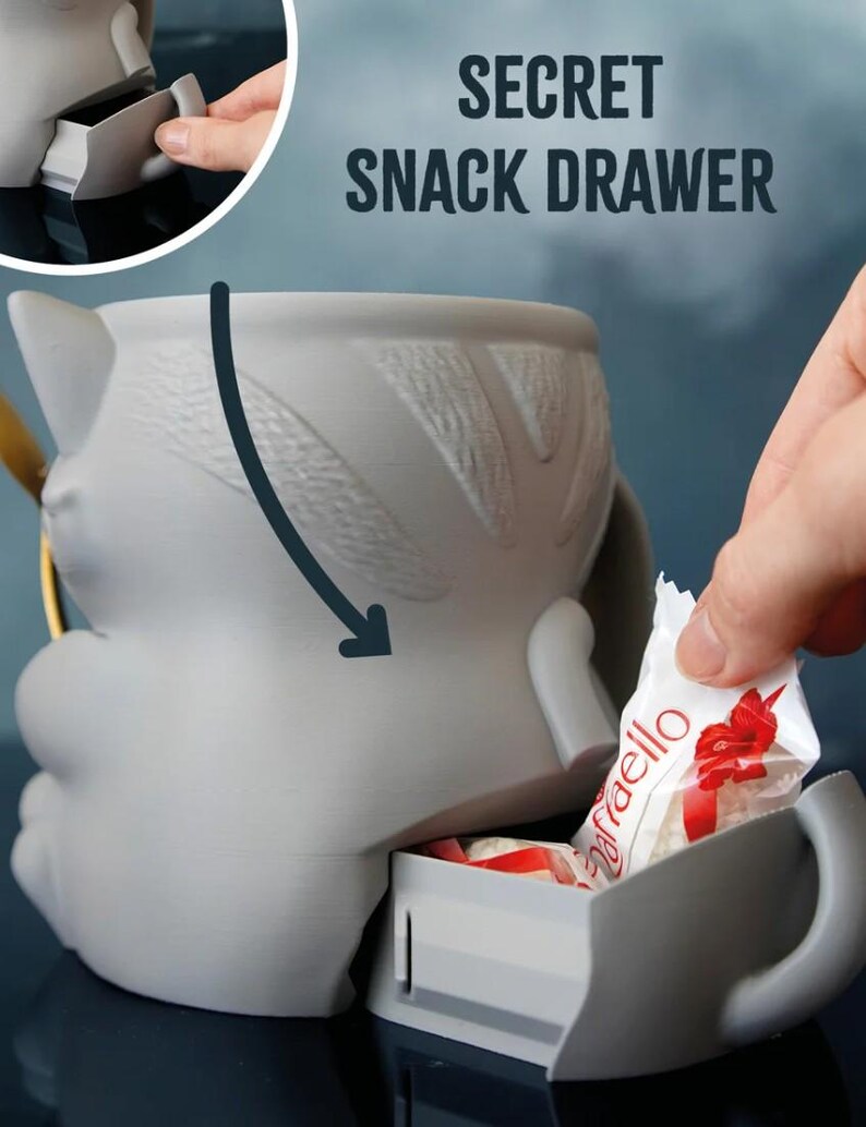 May include: A gray cat-shaped mug with a secret snack drawer. The mug has a handle and a textured design on the back. The words "SECRET SNACK DRAWER" are printed above the mug. A hand is placing a wrapped candy into the drawer.