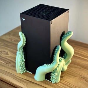 May include: A black gaming console with a green octopus-shaped stand. The console is sitting on a wooden surface.
