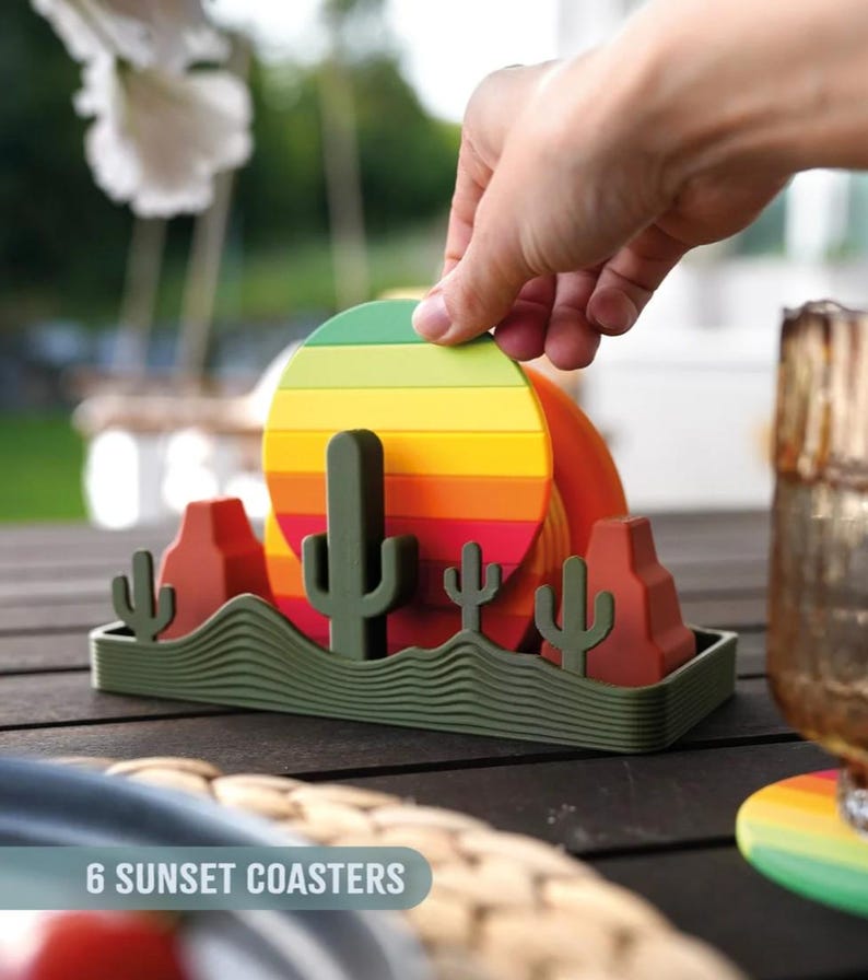 May include: A set of six sunset-themed coasters. Each coaster features a colorful, striped design resembling a sunset. The set includes a green holder with cactus and desert landscape details. The text "6 SUNSET COASTERS" is visible.