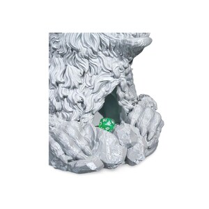 May include: A gray, sculpted dice tower in the shape of a cave entrance. The tower has a textured, rocky exterior and a small opening where a green die with white numbers is visible. The dice tower is designed for tabletop gaming.