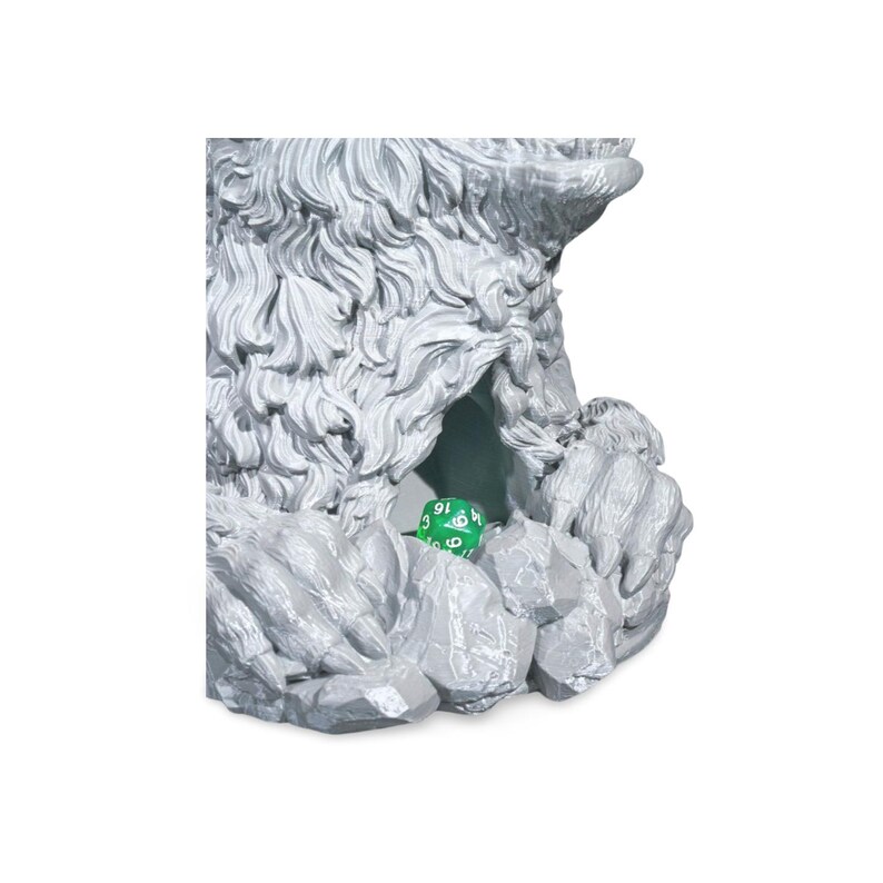 May include: A gray, sculpted dice tower in the shape of a cave entrance. The tower has a textured, rocky exterior and a small opening where a green die with white numbers is visible. The dice tower is designed for tabletop gaming.