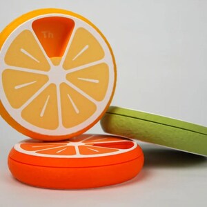 May include: Three pill organizers shaped like citrus fruit slices. One is orange with a white interior, another is orange, and the third is green. The top organizer has the word "Th" printed on it.