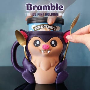 May include: A purple bat-shaped ice cream pint holder with a cartoon face, holding a pint of Ben & Jerry's ice cream. The holder has a handle and two gold spoons. The text "Bramble THE ICE PINT HOLDING BAT" is above the holder.