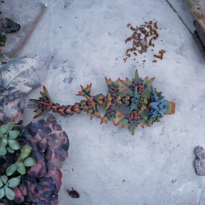 May include: A colorful, intricate, 3D printed dragon sculpture with a green, blue, and orange color scheme. The dragon is lying on a light gray surface.