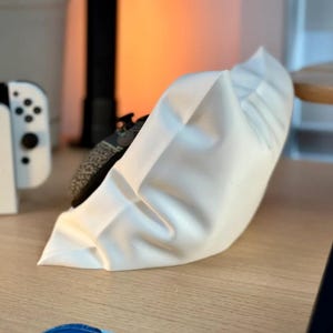 May include: A white, fabric pillow with a ruffled edge. The pillow is sitting on a wooden surface.