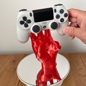 May include: A white PlayStation 4 controller is held by a red 3D printed hand, which is on a white rotating display stand.