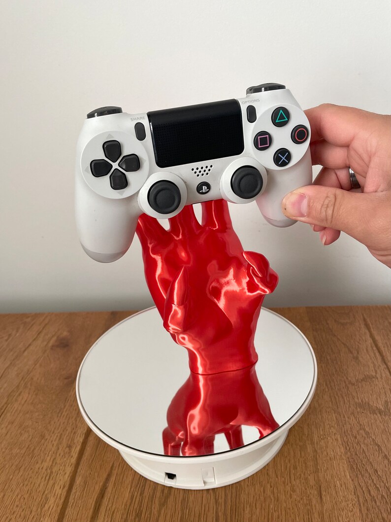 May include: A white PlayStation 4 controller is held by a red 3D printed hand, which is on a white rotating display stand.