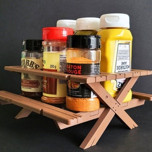 May include: A brown wooden picnic table-style spice rack with five glass spice jars and a yellow mustard bottle. The jars contain various spices, including "Moutarde Jaune", "Piment Rouge", and "Rub BBQ".