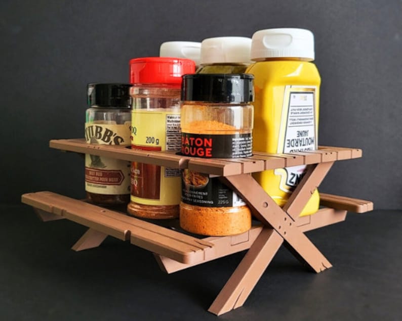 May include: A brown wooden picnic table-style spice rack with five glass spice jars and a yellow mustard bottle. The jars contain various spices, including "Moutarde Jaune", "Piment Rouge", and "Rub BBQ".