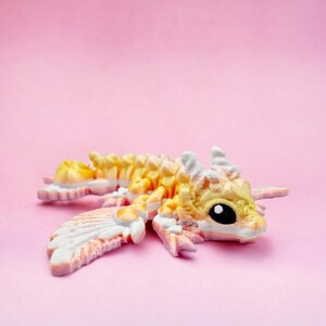 May include: A small, yellow and pink dragon figurine with white wings. The dragon has a black eye and is lying on its side with its wings spread out.