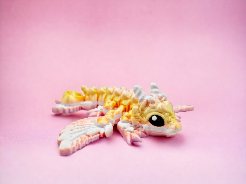 May include: A small, yellow and pink dragon figurine with white wings. The dragon has a black eye and is lying on its side with its wings spread out.