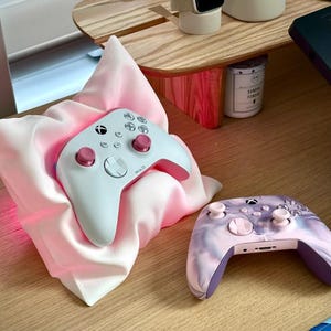 May include: Two white and pink Xbox controllers. One controller is resting on a pink pillow, the other is on a wooden surface. The controller on the pillow has a pink glow around it.