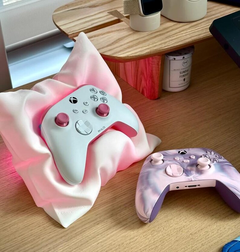 May include: Two white and pink Xbox controllers. One controller is resting on a pink pillow, the other is on a wooden surface. The controller on the pillow has a pink glow around it.