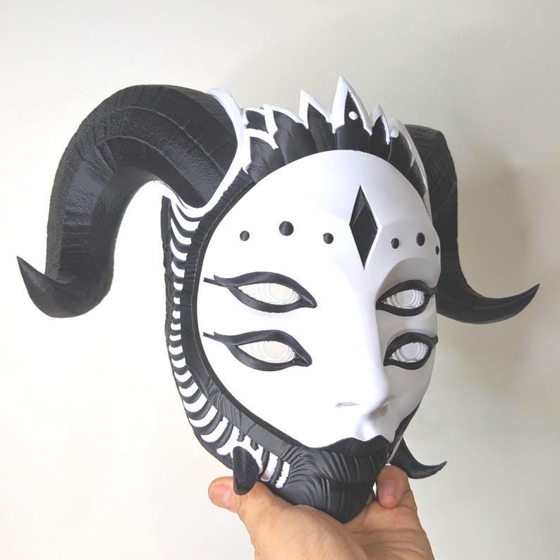 May include: A white and black mask with a horned design. The mask features a face with multiple eyes, a diamond shape on the forehead, and a black beard. The horns are black and curved.