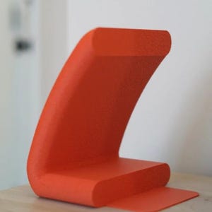 May include: A vibrant orange, abstract sculpture with a textured surface. The design features a curved, wave-like shape rising from a rectangular base, with a small, flat extension. The sculpture is displayed on a light-colored wooden surface.