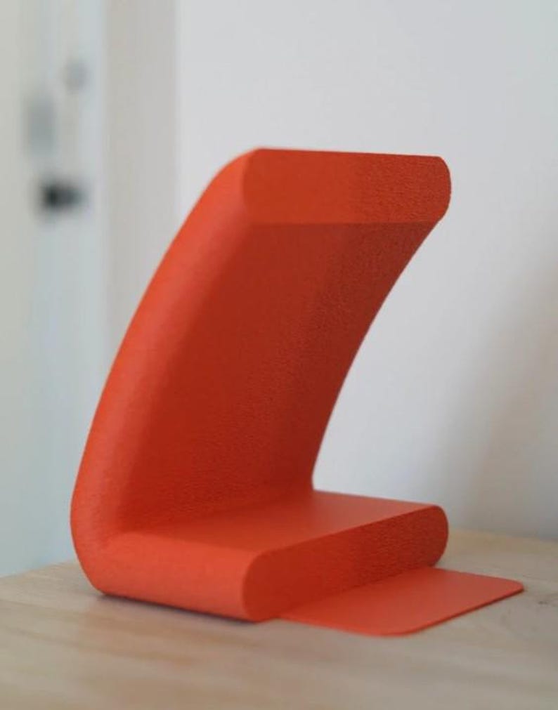 May include: A vibrant orange, abstract sculpture with a textured surface. The design features a curved, wave-like shape rising from a rectangular base, with a small, flat extension. The sculpture is displayed on a light-colored wooden surface.