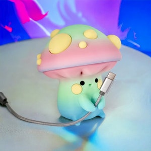 May include: A pastel green and pink mushroom-shaped phone charger with a silver USB-C cable. The charger has a cute cartoon face with yellow eyes and a pink mouth.