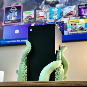 May include: A black Xbox Series X console is held by a 3D printed green octopus stand. The console is sitting on a wooden surface.