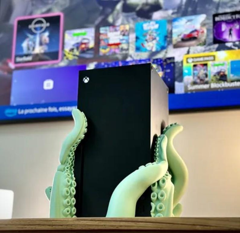 May include: A black Xbox Series X console is held by a 3D printed green octopus stand. The console is sitting on a wooden surface.