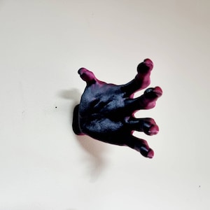 Puede incluir: Una escultura de mano de color morado oscuro y negro con los dedos extendidos, montada en una pared blanca.