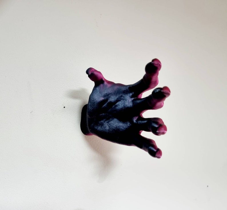 Puede incluir: Una escultura de mano de color morado oscuro y negro con los dedos extendidos, montada en una pared blanca.